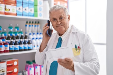 Middle age grey-haired man pharmacist talking on smartphone reading prescription at pharmacy