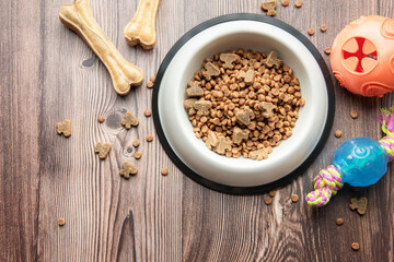 A bowl with dog food, dog treats and toys on a wooden floor.