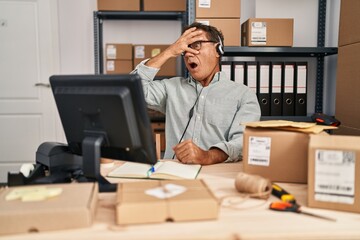 Senior man working at small business ecommerce wearing headset peeking in shock covering face and eyes with hand, looking through fingers afraid