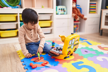 Adorable hispanic boy playing with technician tools toy sitting on floor at kindergarten
