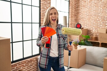 Young blonde woman holding roller painter and paint samples at new home winking looking at the...