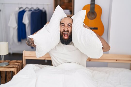 Young bald man covering ears for noise at bedroom