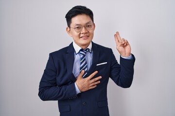 Young asian man wearing business suit and tie smiling swearing with hand on chest and fingers up, making a loyalty promise oath