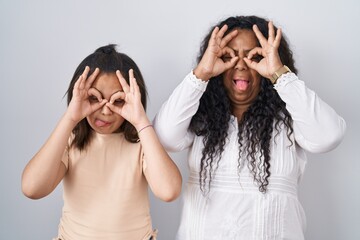 Mother and young daughter standing over white background doing ok gesture like binoculars sticking tongue out, eyes looking through fingers. crazy expression.