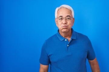 Middle age man with grey hair standing over blue background puffing cheeks with funny face. mouth inflated with air, crazy expression.