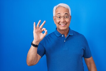 Middle age man with grey hair standing over blue background smiling positive doing ok sign with hand and fingers. successful expression.
