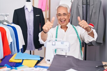 Middle age man with grey hair dressmaker using sewing machine approving doing positive gesture with hand, thumbs up smiling and happy for success. winner gesture.