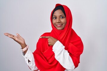 Young arab woman wearing traditional islamic hijab scarf amazed and smiling to the camera while presenting with hand and pointing with finger.