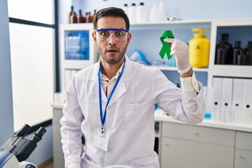 Young hispanic man with beard working at scientist laboratory holding green ribbon scared and amazed with open mouth for surprise, disbelief face