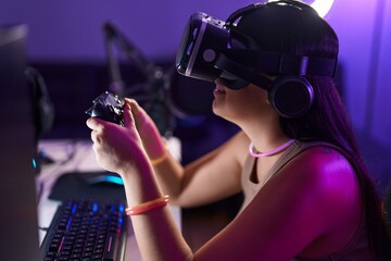 Young beautiful hispanic woman streamer playing video game using virtual reality glasses and joystick at gaming room