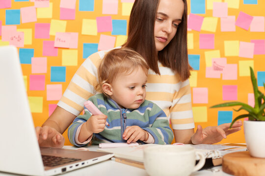 Portrait of serious dark haired woman accountant wearing striped t-shirt sitting at table, posing against memo cards on yellow wall, playing with baby and using calculator.