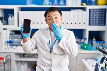 Young chinese man working at scientist laboratory holding smartphone covering mouth with hand,...