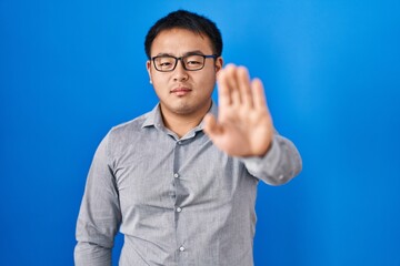 Young chinese man standing over blue background doing stop sing with palm of the hand. warning...