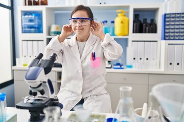 Hispanic girl with down syndrome working at scientist laboratory smiling pulling ears with fingers, funny gesture. audition problem