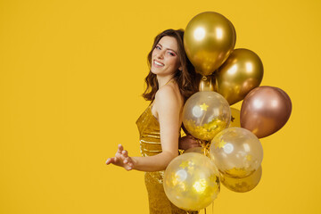 Fototapeta premium Festive concept. Charming lady in dress with bunch of air balloons having holiday party, yellow background, copy space