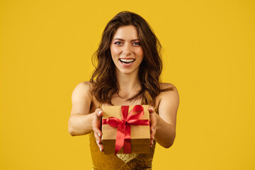 Holidays celebration. Happy beautiful lady in dress showing wrapped box with gift, posing on yellow background