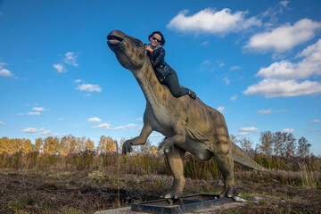 Naklejka premium Beautiful girl near the dinosaur. Girl in a leather jacket. Sunny day. Dinosaur in the field.