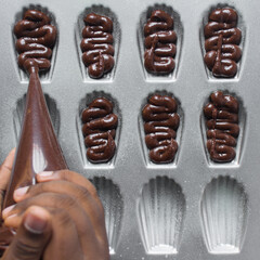 Piping chocolate Madeleine batter into a greased Madeleine pan, chocolate cake batter in a baking pan