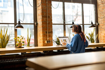 Young stylish woman works on laptop while sitting by the big window at modern coffee shop. Wide view from the backside
