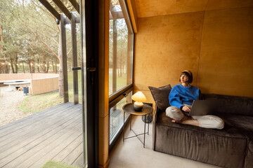 Woman works on laptop while sitting relaxed on a couch in wooden house in forest. Concept of remote work from cozy house on nature