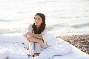 Smiling young woman wear silk pajamas sitting in bed with white duvet and pillows over sea shore...