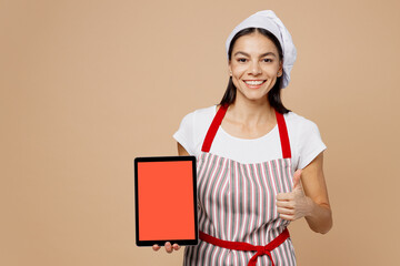 Young housewife housekeeper chef baker latin woman wear striped apron toque hat hold use digital tablet pc computer show recipe show thumbup isolated on plain light beige background Cook food concept