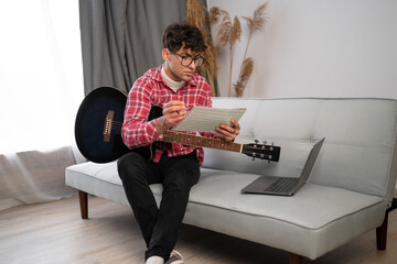 Young arabic composer man composing song using guitar and laptop at home sitting on sofa.