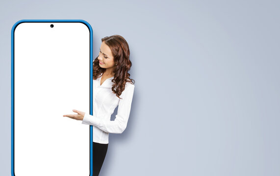 Smiling Business Woman, Showing Big Cell Phone, Mobile Smartphone. Happy Businesswoman Holding Peeping Out From Cellphone With White Mockup Screen, Isolated Over Grey Background. App Ad Concept Image.