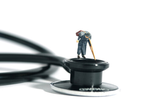 Miniature people toy figure photography. A old man with stick or crutch standing above stethoscope. Medical checkup concept