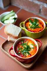 Two portions of shakshuka in red and green baking dishes on a wooden tray.  Traditional Israeli and Arabic dish shakshuka consisting of eggs poached in a spicy sauce of tomatoes, onion, bell