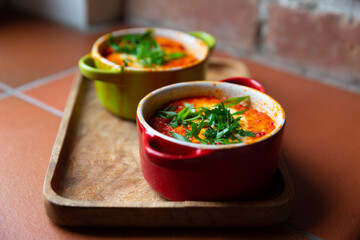 Two portions of shakshuka in red and green baking dishes on a wooden tray, vertical.  Traditional Israeli and Arabic dish shakshuka consisting of eggs poached in a spicy sauce of tomatoes, onion, bell