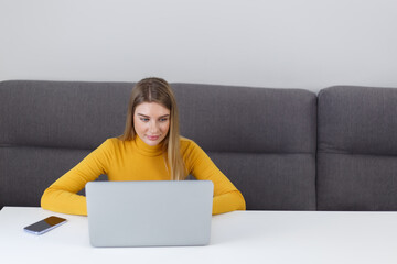 Young student girl studing on laptop computer at home. Beautiful blonde female person sitting at the table and working on modern notebook pc