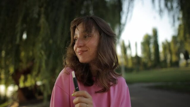Cheerful young girl in a pink t-shirt in the park smokes an electronic cigarette. Cute girl in a pink t-shirt, smokes an electronic cigarette in the park. A beautiful young woman with an e-cigarette.