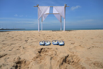 Two pairs of white flip-flops with blue ears lined up on the sandy beach fronted by bamboo arches decorated with white curtains on a summer's day.White bamboo arches installed on the beach for tourist