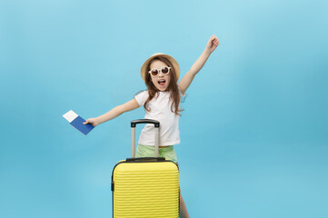 Cute little girl holding boarding pass, expressing happiness at camera, standing with yellow suitcase on blue background