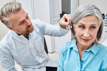 Audiologist fits hearing aid on deafness mature woman ear during visit to audiology center. Hearing...