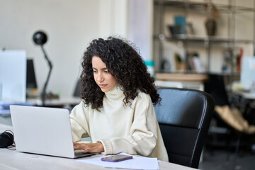 Young latin professional business woman worker sitting at desk working on marketing data online on laptop in modern corporate office. Female worker using computer technology typing browsing web.
