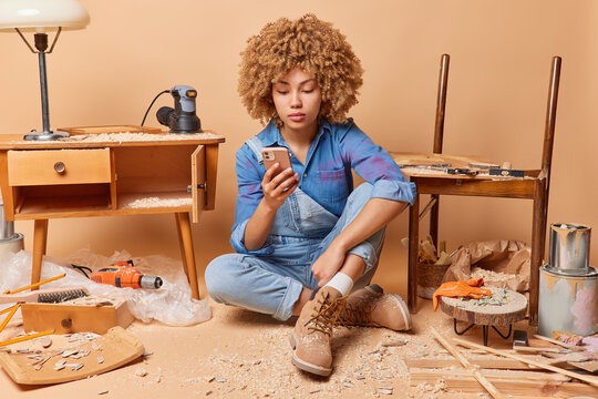 Serious feamale joiner dressed in comfortable clothes uses mobile phone checks received messages produces wooden furniture involved in woodworking sits on floor in messy room beige background
