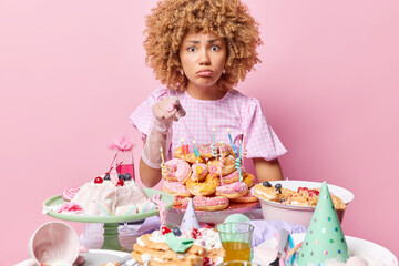 Indoor shot of curly woman points at you with reproach feels sad poses near festive table with desserts cone hats celebrates birthday isolated over pink background. Festive event celebration concept
