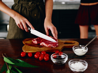 Woman with knife in hand cutting steak meat for frying in kitchen with salt pepper and other spices on table, red cherry tomatoes and herbs, dinner preparation.