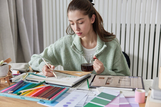Professional female designer or artist draws sketches works on design of apartment poses at desktop with prepares interior design drinks tea wears casual jumper. Color samples for selection.