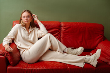 Fashionable confident woman wearing elegant white knit suit, with zip neck sweater, wide leg trousers, leather ankle lace up boots, sitting, posing on leather couch. Copy, empty space for text - 565953144