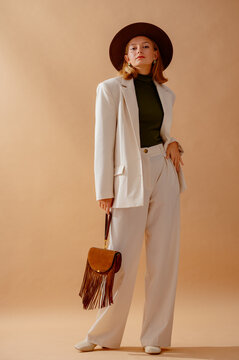 Fashionable confident woman wearing elegant white suit with blazer, wide leg trousers, turtleneck top, hat, leather ankle boots, holding brown suede fringed bag,  posing on beige background