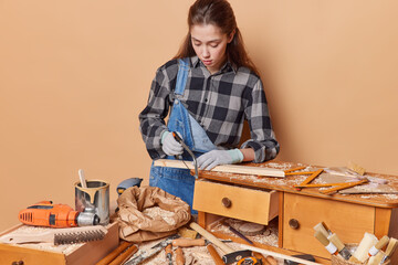 Fototapeta na wymiar Indoor shot of female carpenter works making woodcraft furniture cuts wooden plank with jigsaw wears checkered shirt denim overalls and overalls fixes table uses different instruments for manual work