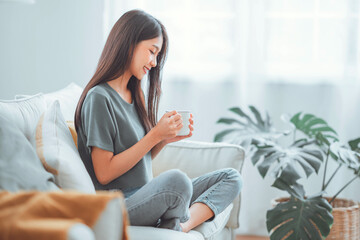 Happy young asian woman drinking coffee or tea relaxing on sofa at home. Smiling female enjoying resting sitting on couch in modern living room.