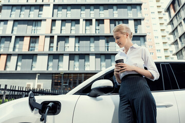 Businesswoman drinking coffee, leaning on electric vehicle recharging at public charging station...
