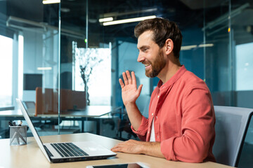 Portrait of a young man in a red shirt. Sitting in the office at the table, working on a laptop, communicating via video call, waving, smiling.