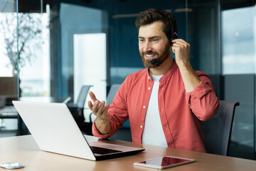 Portrait of a young man in a headset and a red shirt. He sits at the desk in the office, works on a laptop, communicates through a video call, talks, smiles.