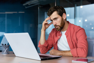 Fototapeta na wymiar A stressed and tired young businessman, student, freelancer in a red shirt sits in the office at a laptop. holding his head. Overtime work, deadline, problems.