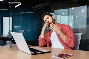 A tired young man in a red shirt sits at the desk in the office and yawns, covering his mouth with his hand. Works overtime, boring monotonous work, falls asleep.